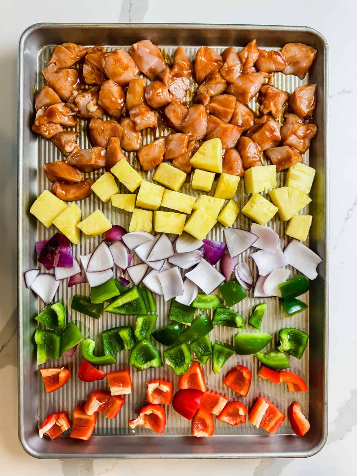 This is a top view of the Hawaiian sheet pan chicken on an XL baking sheet before it is cooked. It features all of the ingredients separated by layer. Chicken, pineapple. red onion, green pepper and red peppers. 